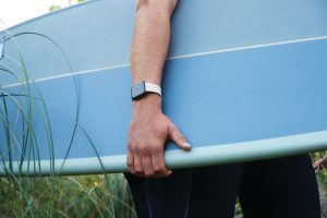 Lifestyle photo of young male holding surfboard, close up of arm wearing product